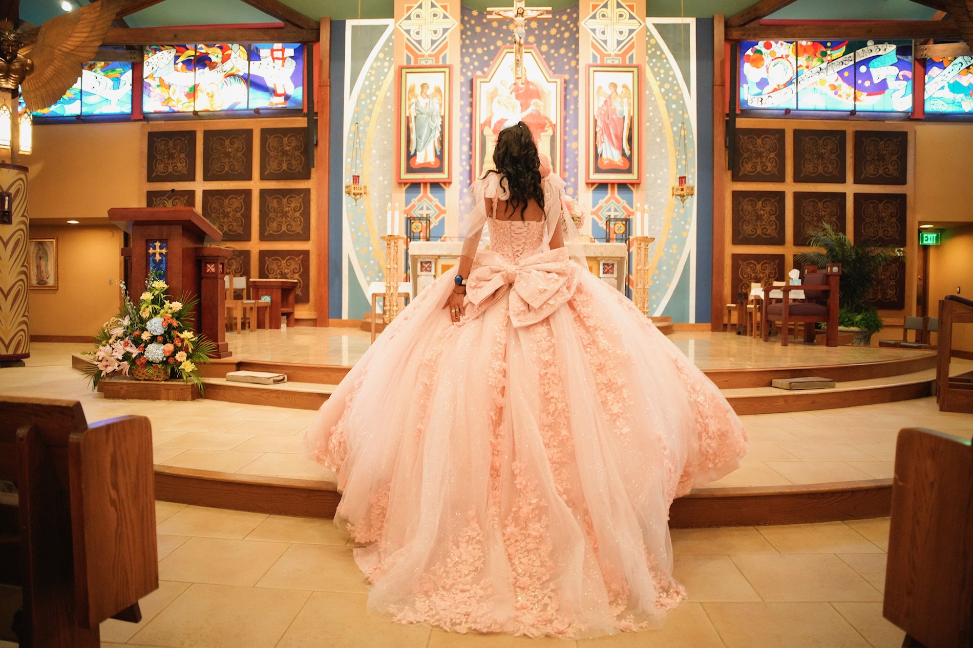 A woman in a pink, elaborate gown with a large bow at the back, standing in a church or chapel. She is facing an altar with religious artwork and a crucifix overhead. There are stained glass windows and floral arrangements in the background.