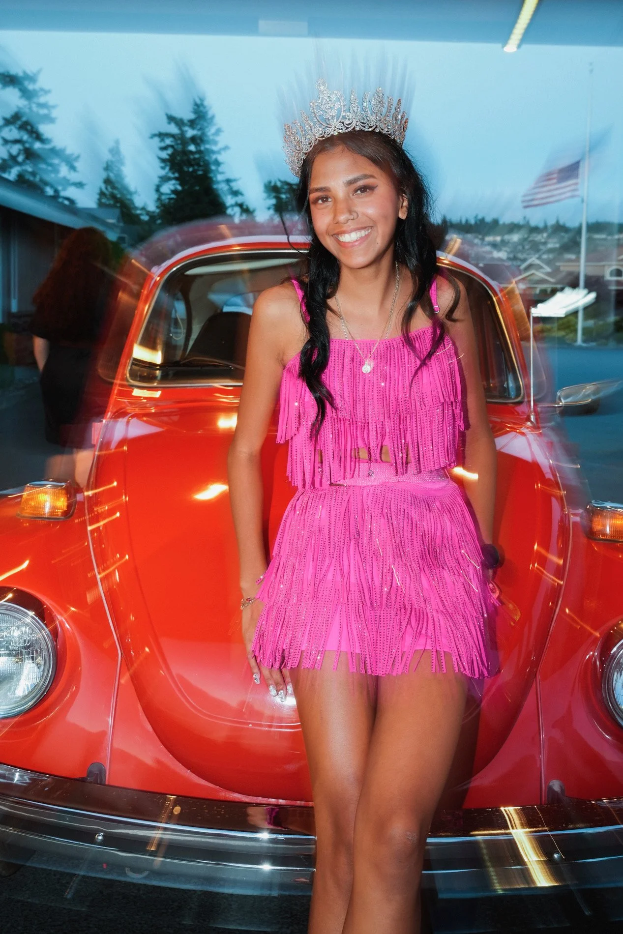 A young woman in a bright pink fringed dress and a silver crown, smiling and standing in front of a red vintage car with a background of trees and a blue sky.