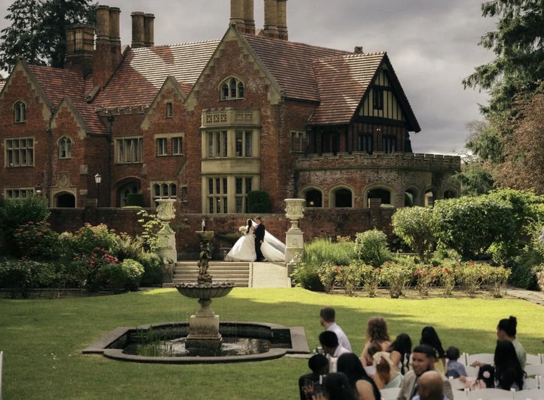 A wedding ceremony taking place outdoors on the lawn of a large, historic brick mansion with elaborate landscaping, a fountain in the foreground, and guests seated watching the bride and groom at the top of stairs.
