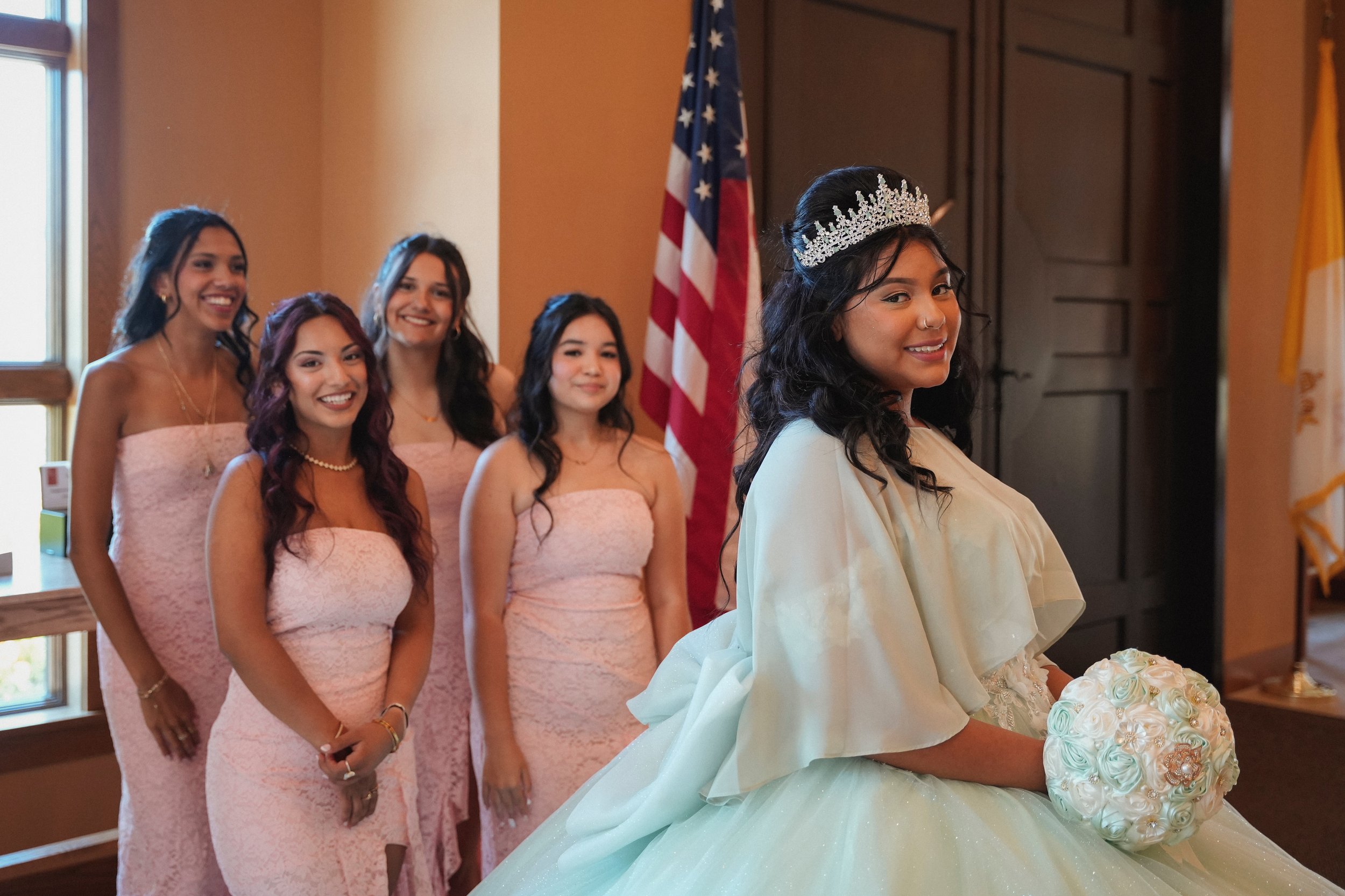 A bride holding a bouquet of white roses stands in front of five women dressed in pink dresses, smiling, with American flags in the background.