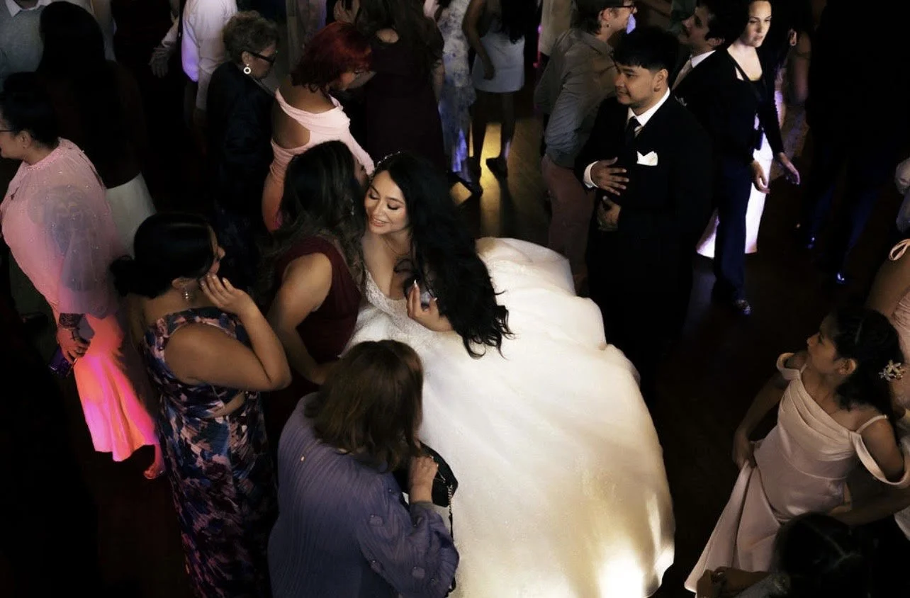 A bride in a white wedding dress is sitting on a table at a wedding reception, surrounded by guests. Guests are standing around, chatting and taking photos in a dimly lit room.