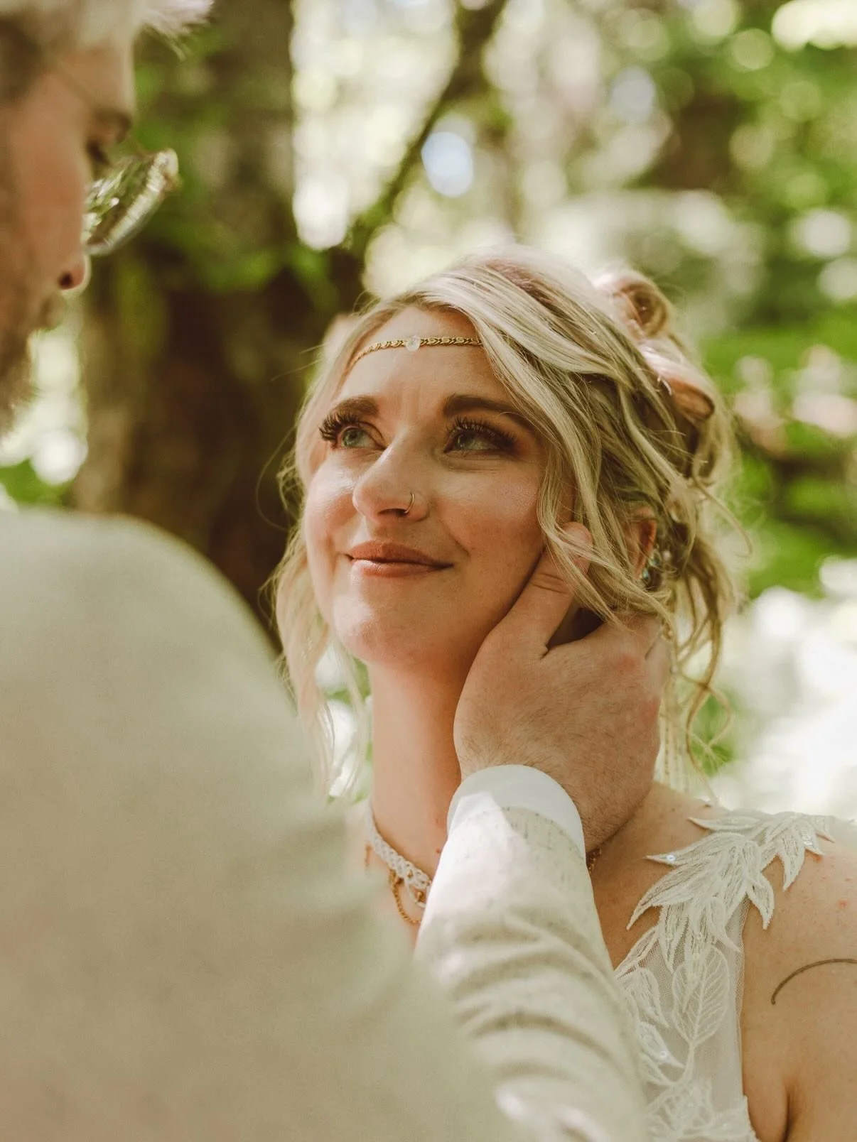 A woman with blonde hair and a gentle smile looking at a man, with his hand tenderly holding her face, in a natural outdoor setting with dappled sunlight and foliage in the background.