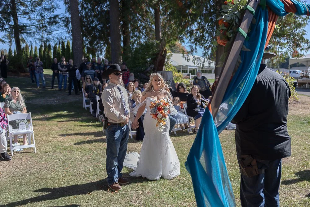 A bride in a white wedding dress holding a bouquet stands next to a man in a cowboy hat and sunglasses at an outdoor wedding ceremony. Guests are seated in chairs and standing behind in a grassy area under trees with blue sky overhead.