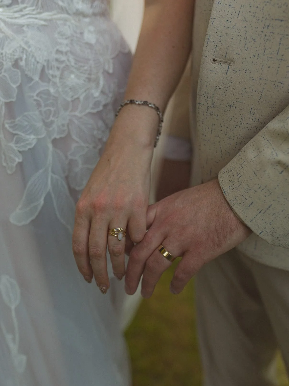 Close-up of a bride and groom holding hands, showing wedding rings. The bride's hand has a gold ring with a gemstone, and she wears a bracelet and a lace wedding dress. The groom has a gold wedding band and is dressed in a light-colored suit.