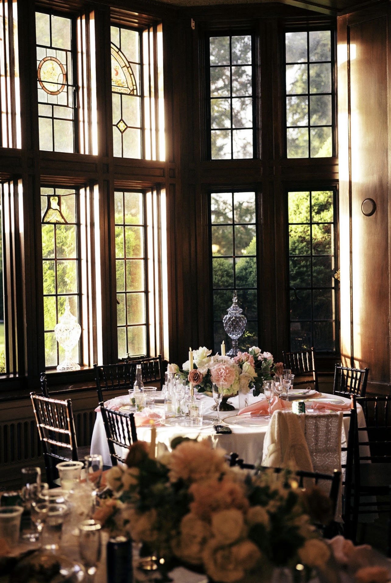 Elegant indoor dining area with large stained glass windows, a round table set with white tablecloth, floral centerpiece, and dinnerware, illuminated by natural sunlight.