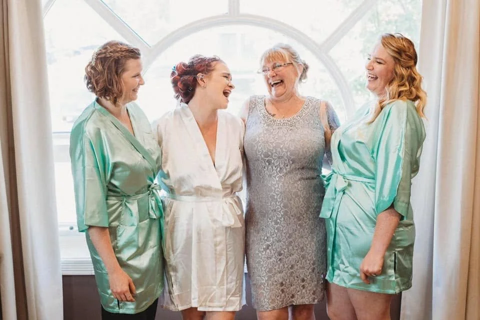 Four women, two in satin robes and two in lace dresses, laughing and standing together in front of a window with curtains on either side.