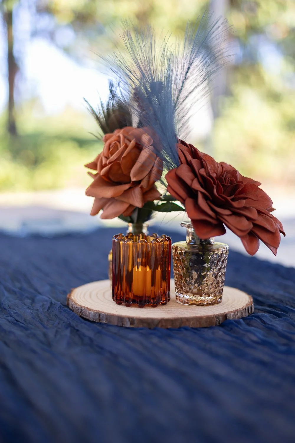 Two small glass vases with artificial orange roses and feathers on a wooden coaster.