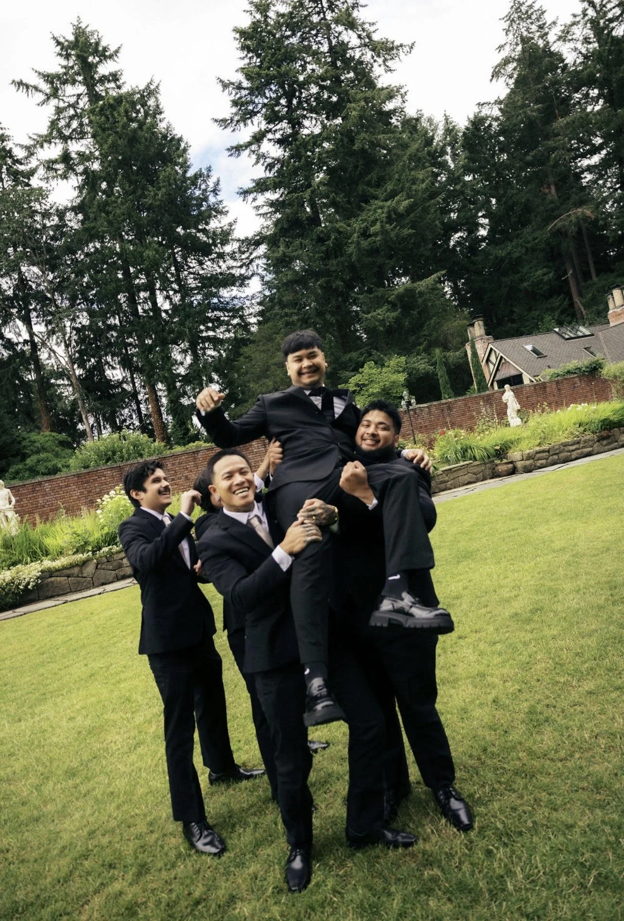 A group of five men dressed in formal suits outdoors in a garden, with one man being lifted by the others. They are smiling and appear to be celebrating.