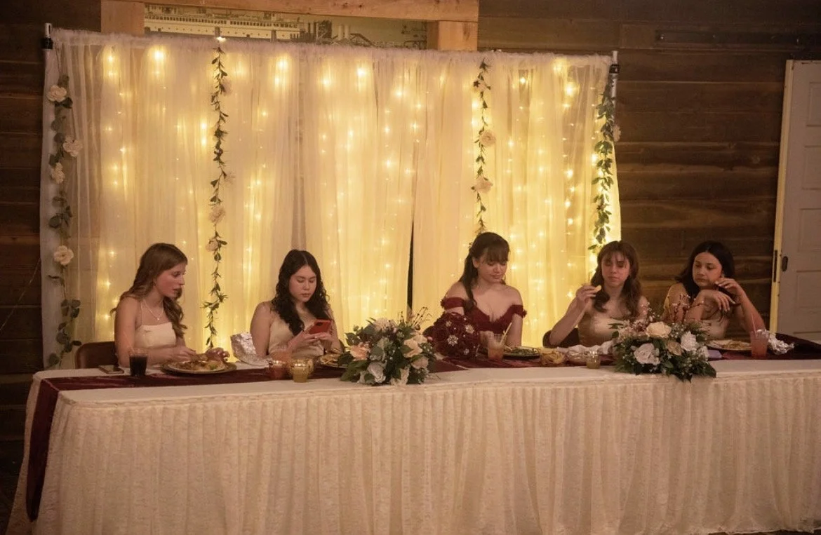 Five young women sitting at a decorated head table, likely at a celebration or wedding reception, with a backdrop of twinkling fairy lights and floral arrangements.