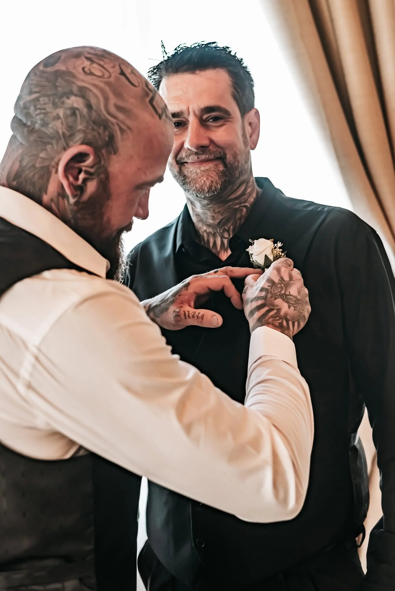 Two men, both with tattoos, are interacting closely indoors. One man, with a beard and dark hair, is smiling gently while clutching a white flower to the other man's chest. The other man, who is bald and heavily tattooed, is dressed in a light-colore