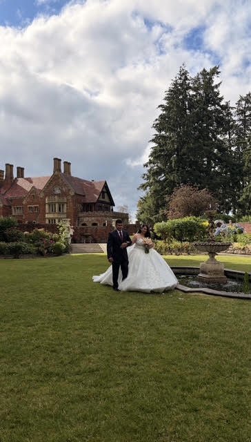 A bride and groom walking on a lawn near a fountain in a large garden, with a historic brick mansion and trees in the background on a partly cloudy day.