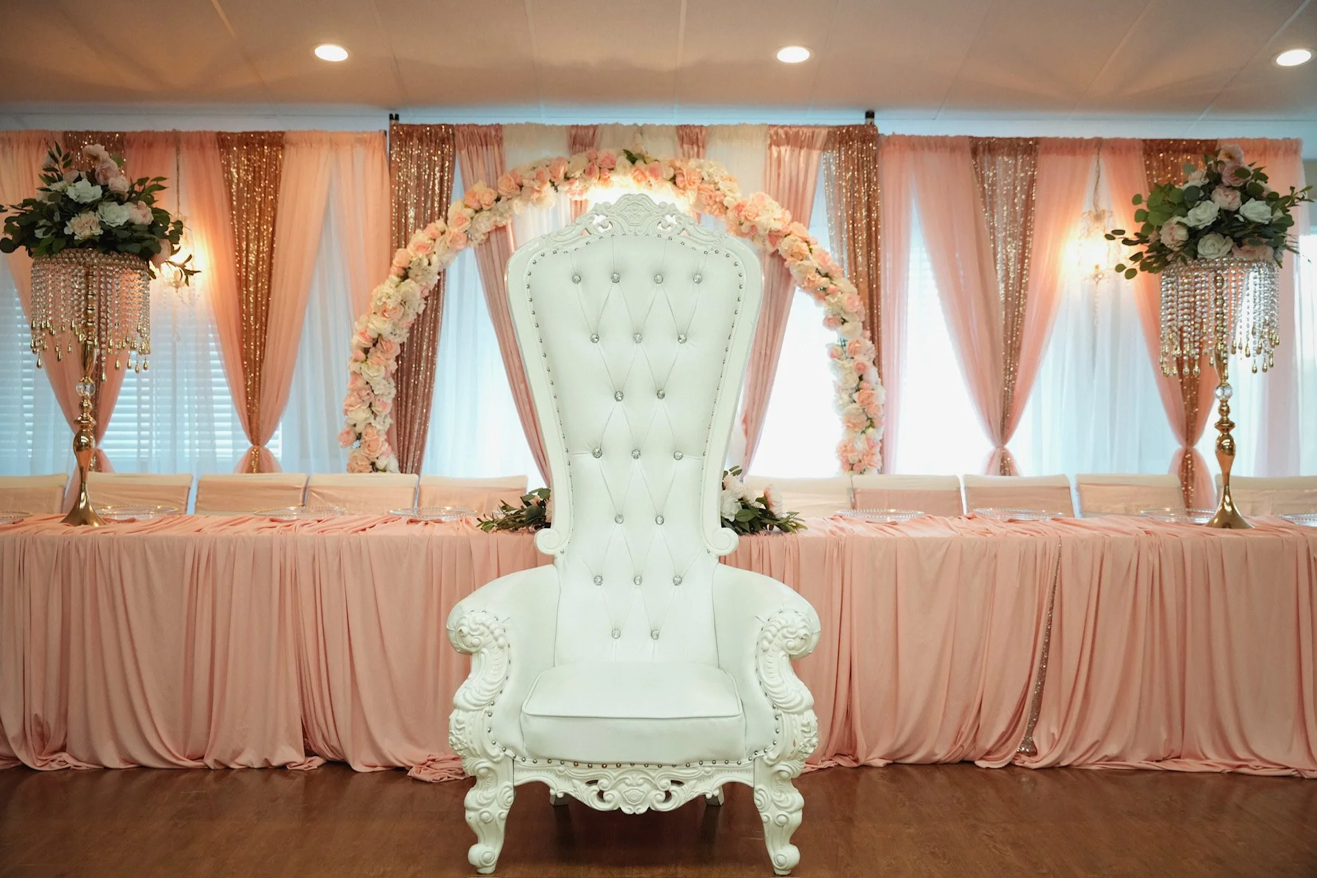Elegant event table setup with a high-backed white throne in front, surrounded by pink drapes, floral arrangements, and gold accents.
