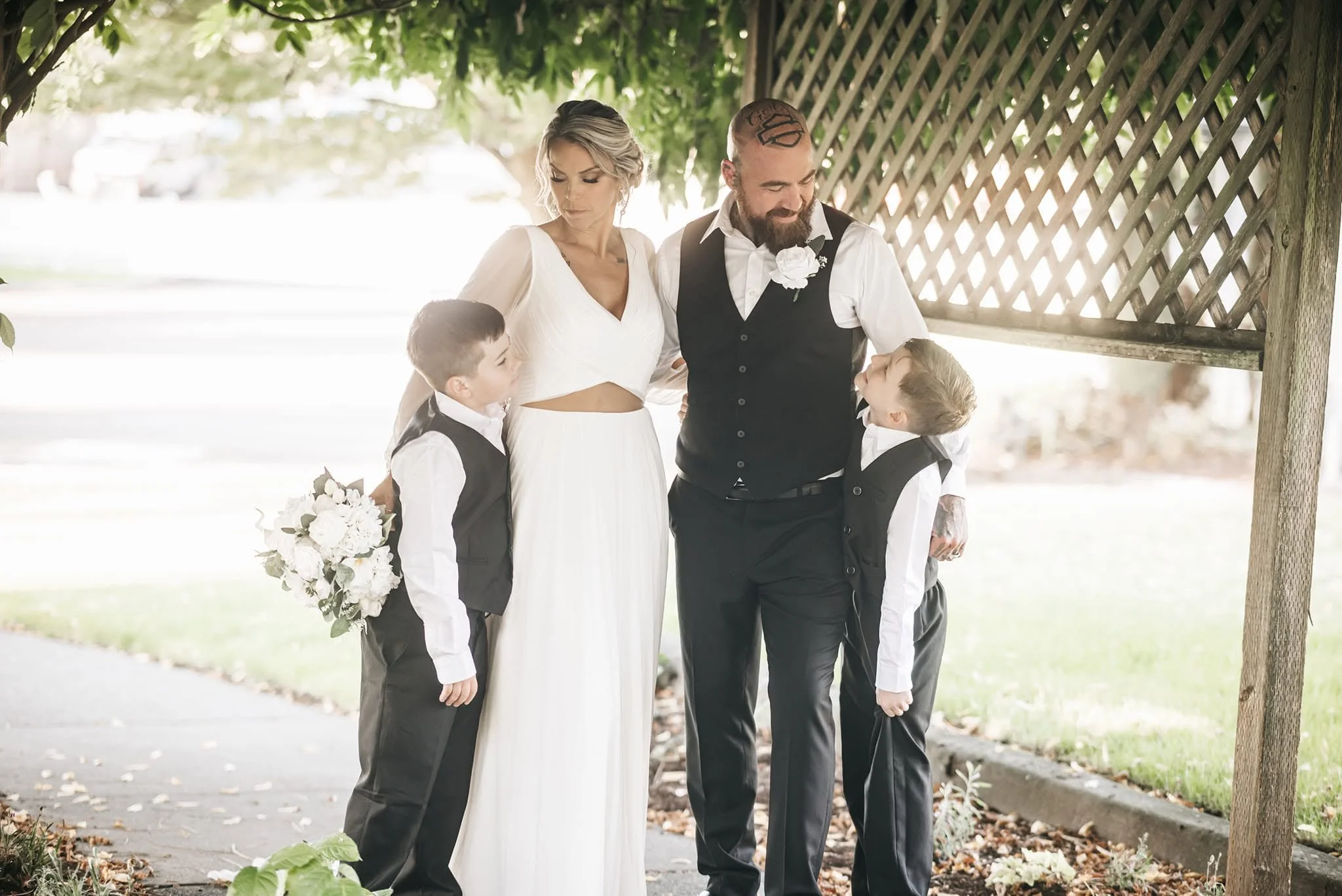 A wedding scene outdoors under a tree with four people, including a woman in a white dress and three men in formal attire, two children and an adult man, sharing a moment together.