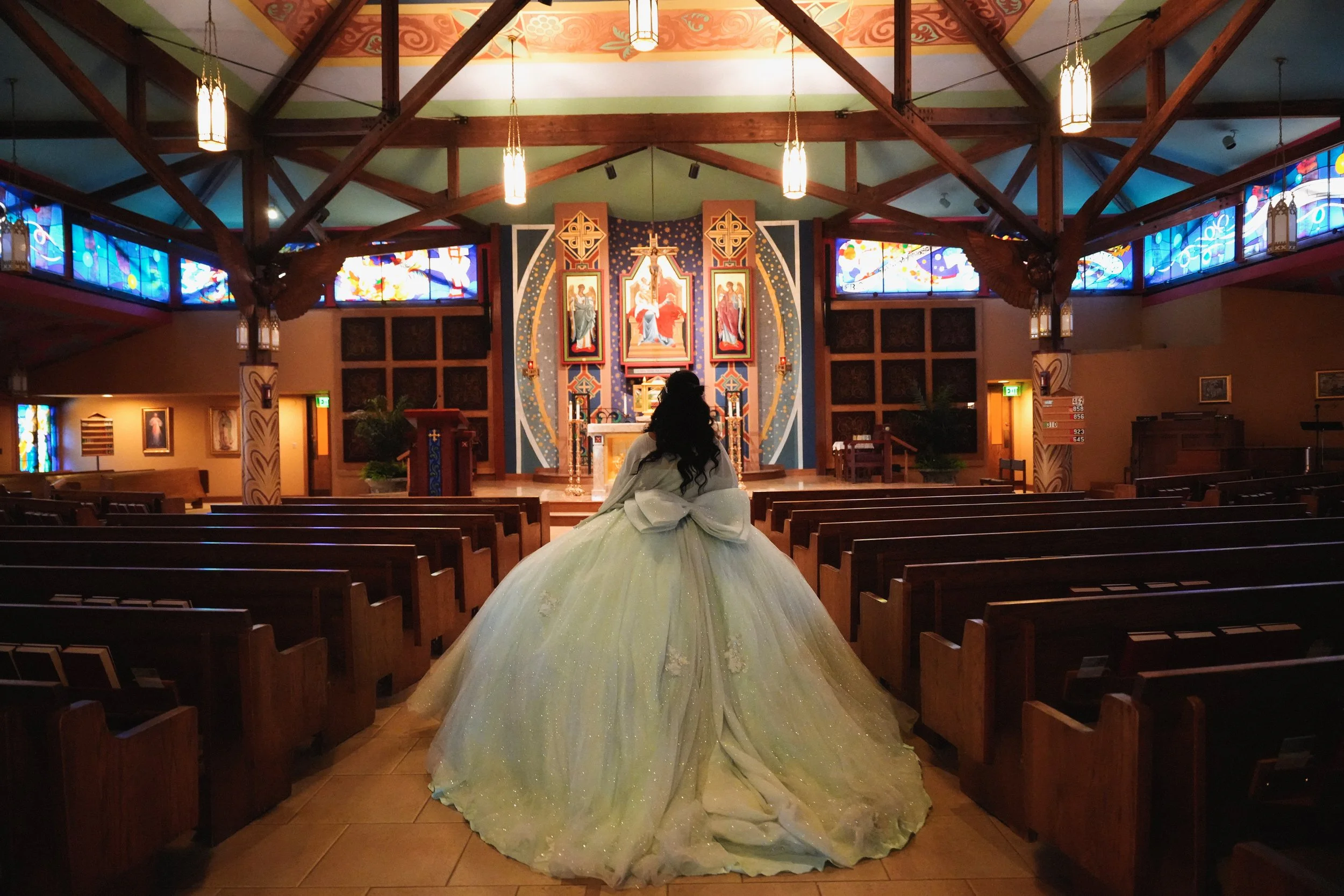 A woman in a large, sparkly, pale green ball gown with a long train sitting inside a church or chapel, facing the altar.