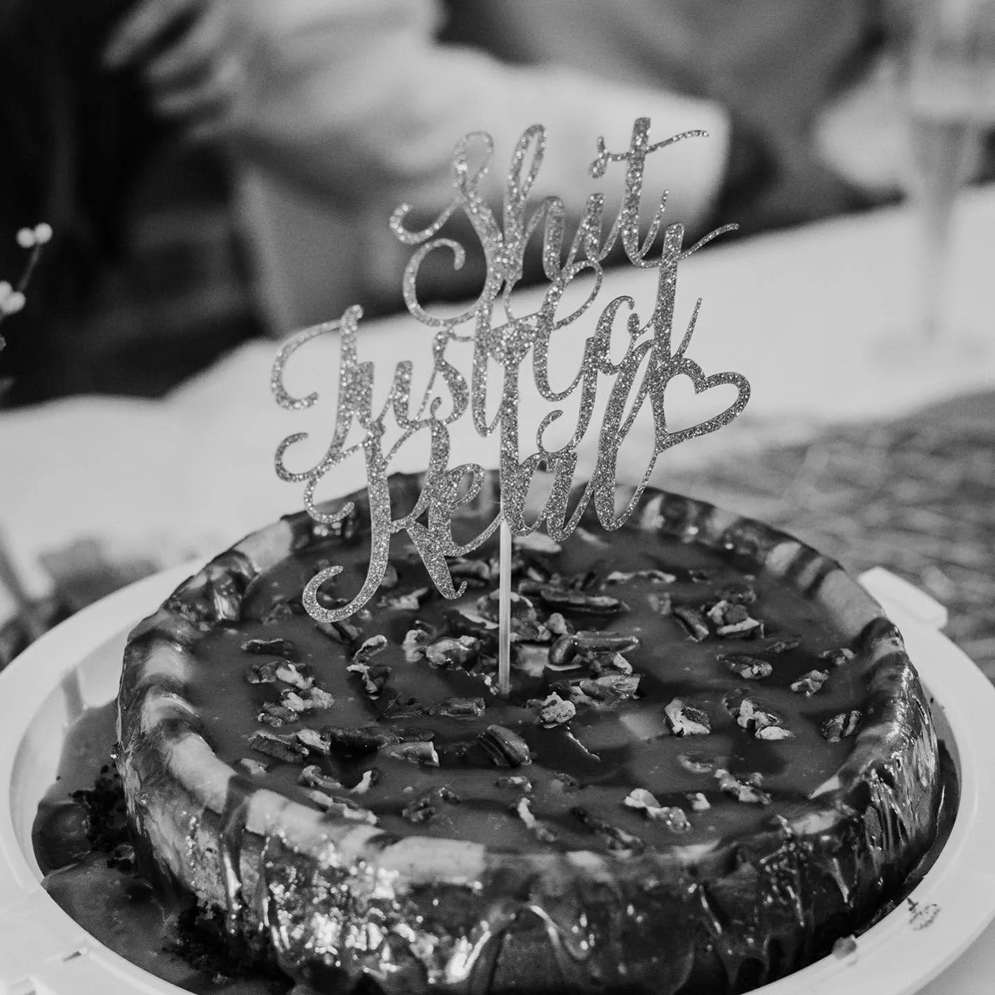 A birthday cake topped with a glittery 'Happy Birthday' sign, decorated with pecans or similar nuts around the edge, placed on a white plate.