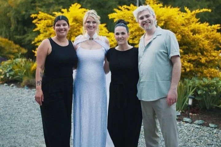 Four people standing outdoors in front of yellow flowering bushes, smiling at the camera. The group includes two women in black dresses, one woman in a light-colored dress, and a man in a light-colored shirt and pants.