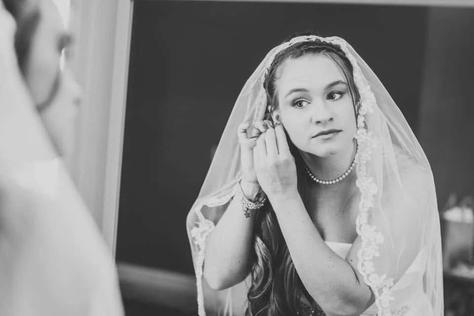 Bride wearing a veil and pearl necklace, adjusting her earring in front of a mirror.