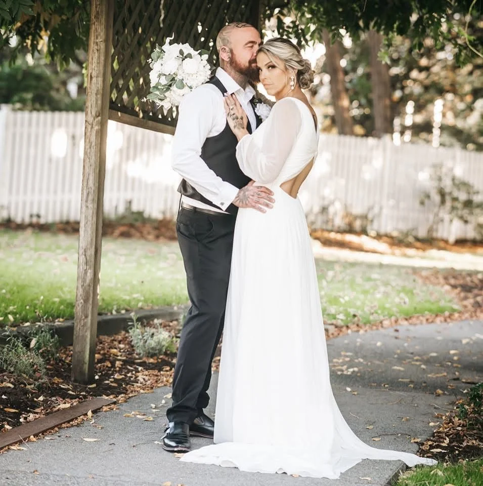 A couple in wedding attire poses closely outdoors near a wooden arbor with flowers, in front of a white picket fence and green trees.