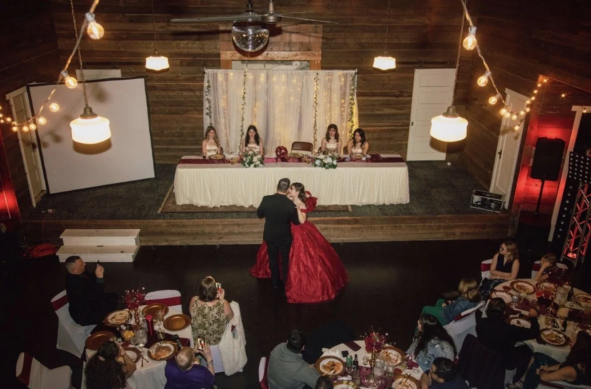 A couple is dancing at a wedding reception with a sweetheart table in the background and guests watching.
