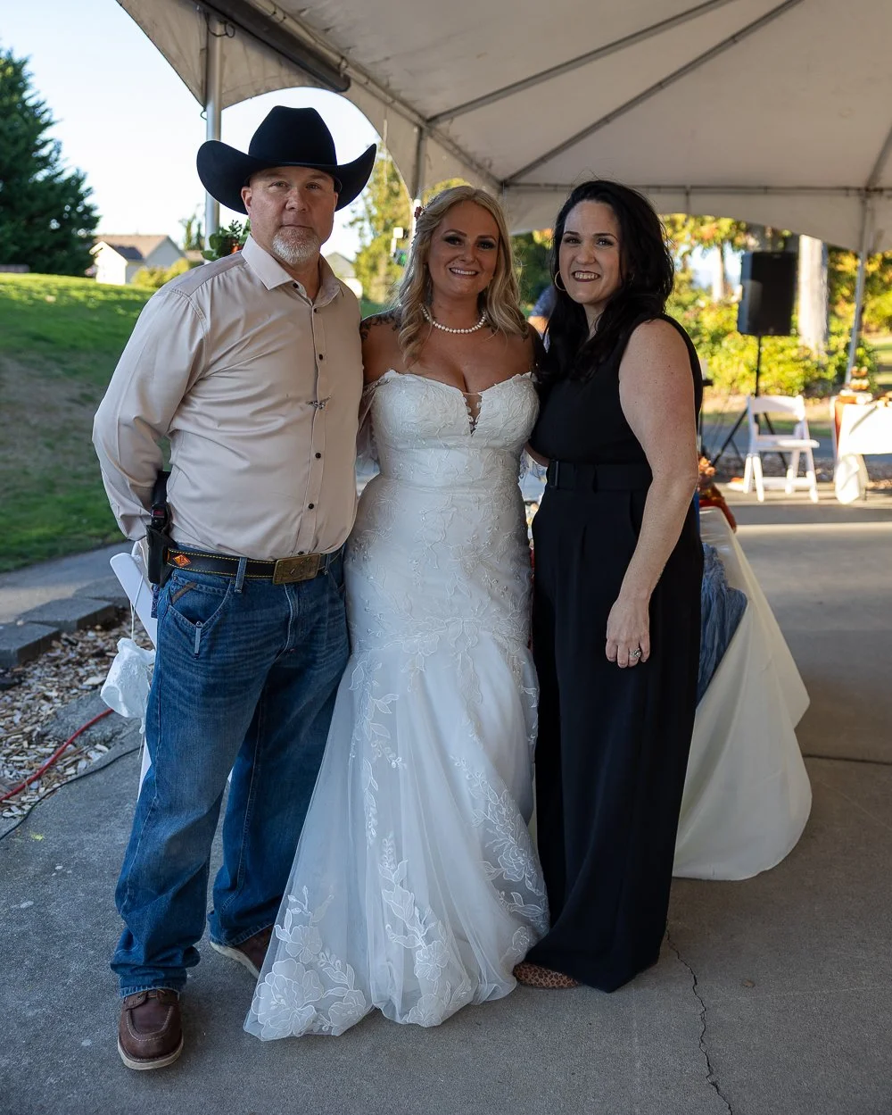 Three people standing under a tent at a wedding reception, with a man in cowboy hat, a bride in a white wedding dress, and a woman in a black dress.