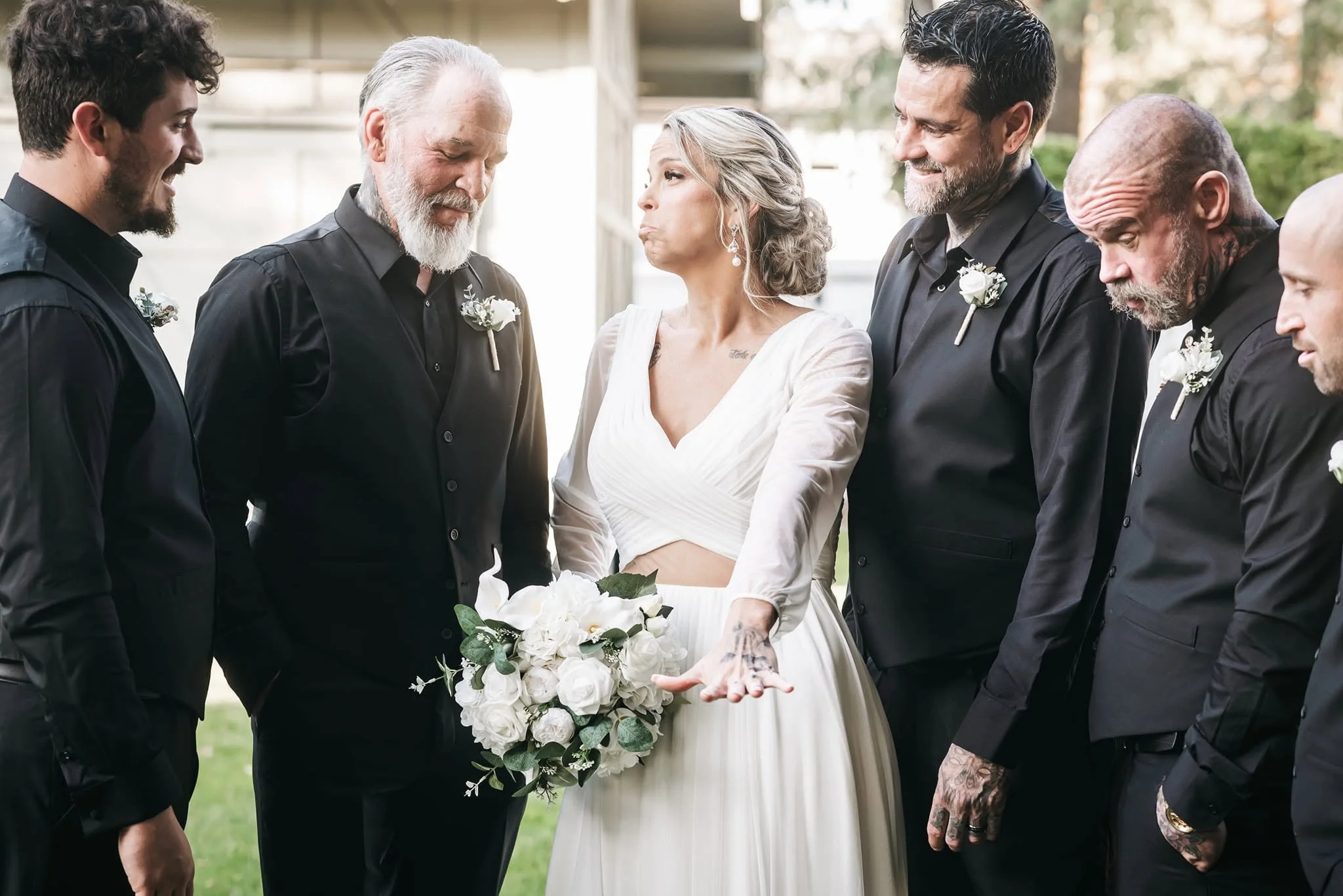 A bride in a white wedding dress holding a bouquet of white flowers and greenery, standing in the center with five men dressed in black around her, all smiling. The men have various hairstyles and beards, and some wear white boutonnieres. The scene i