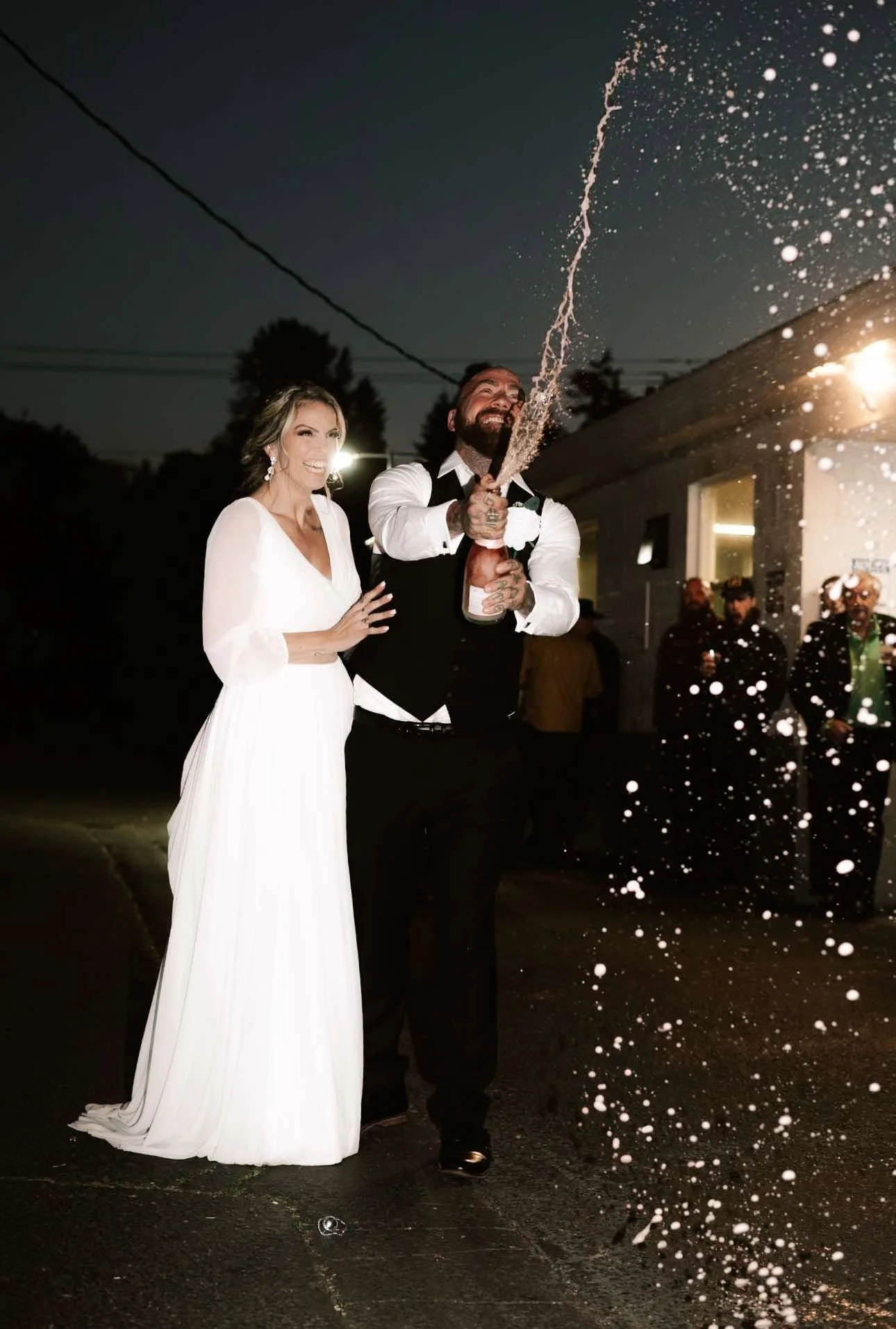 A newlywed couple celebrating outdoors at night, with the groom opening a champagne bottle, spraying it into the air. The bride is wearing a white wedding dress, and the groom is dressed in a black vest and white shirt. They are smiling and surrounde