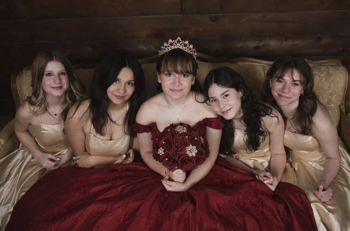 A bride in a red gown with a tiara sitting on a sofa, holding a bouquet of red roses, surrounded by her bridesmaids in gold dresses.