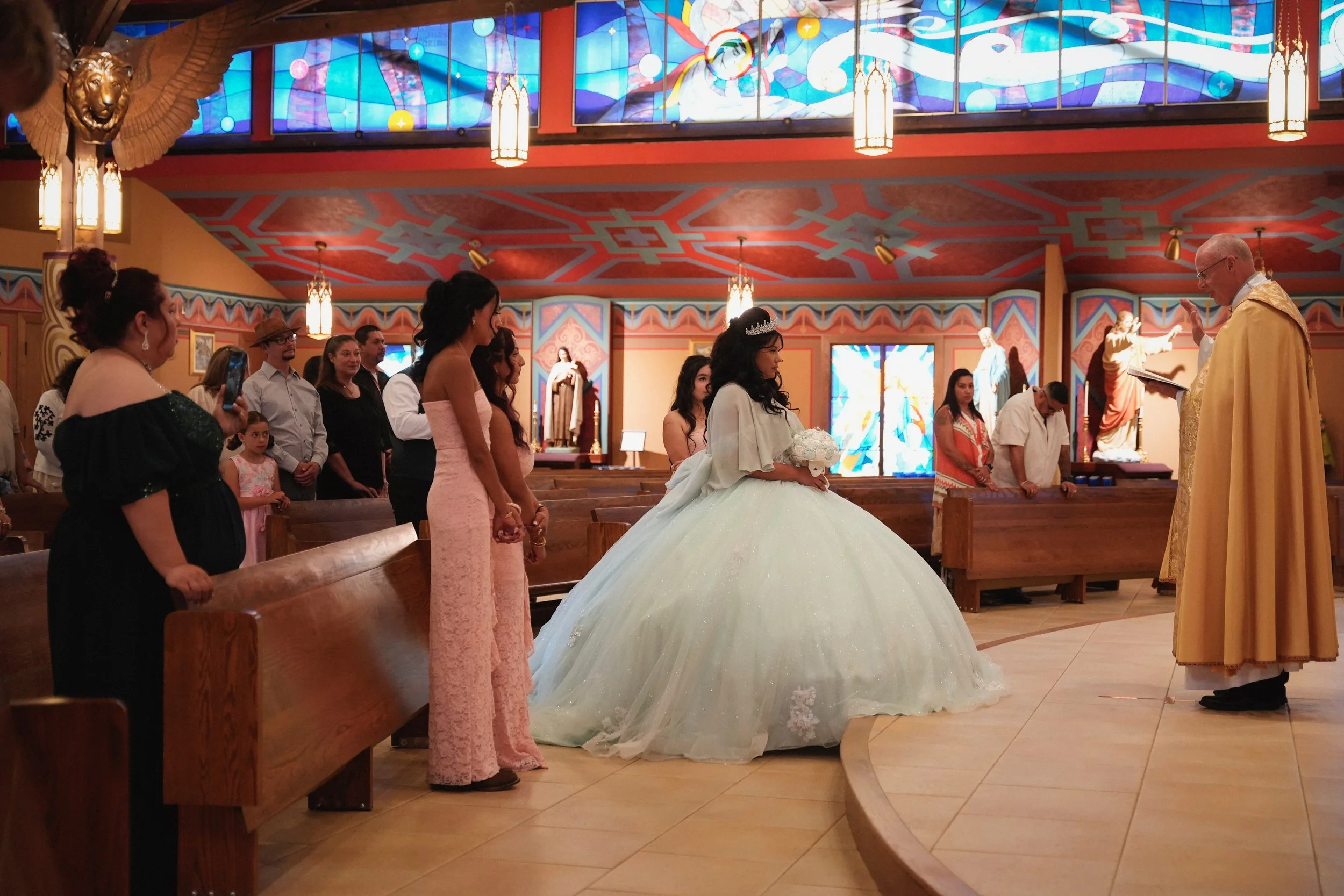 A bride in a white wedding gown and tiara sits in a church, surrounded by people, with a priest and guests participating in a wedding ceremony. The church has colorful stained glass windows and ornate decorations.