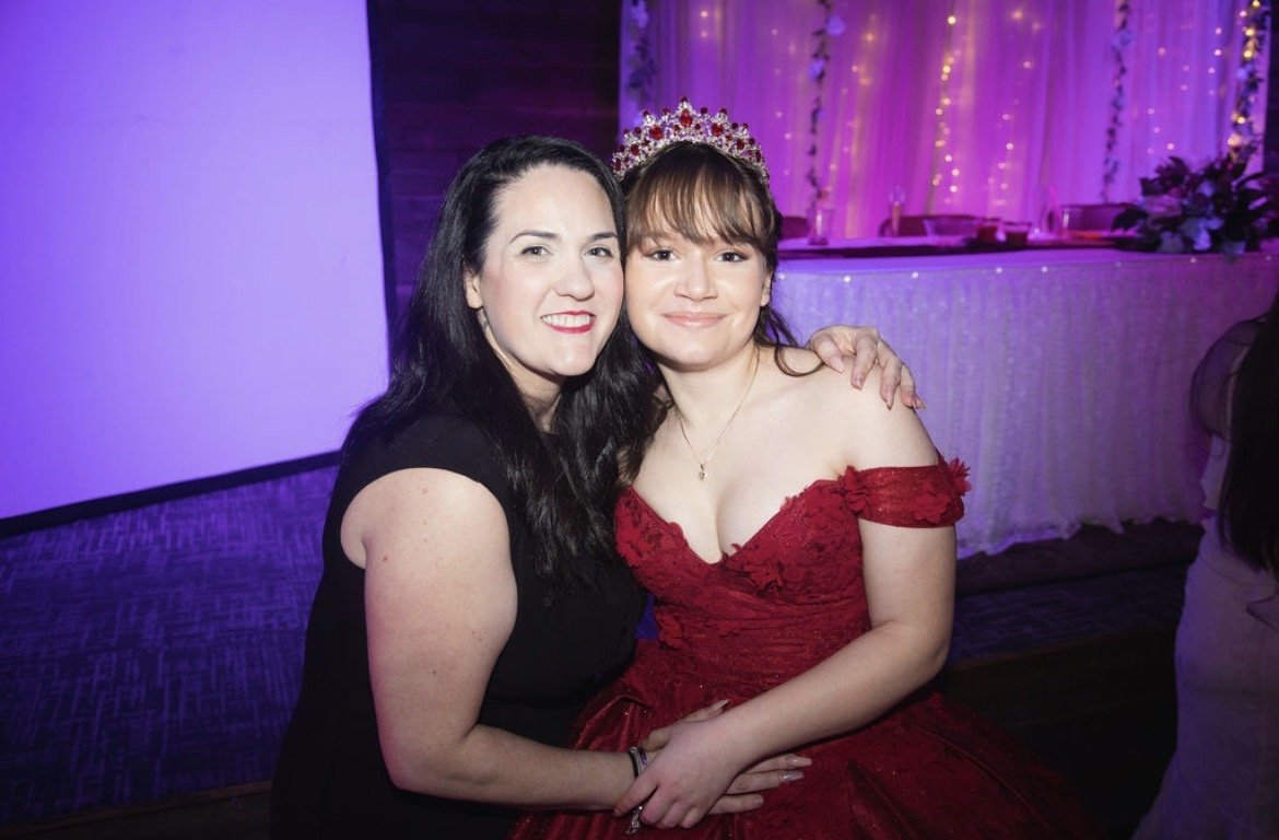 Two women at a celebration; one in a black dress and the other in a red gown with a tiara, with purple and pink lighting in the background.