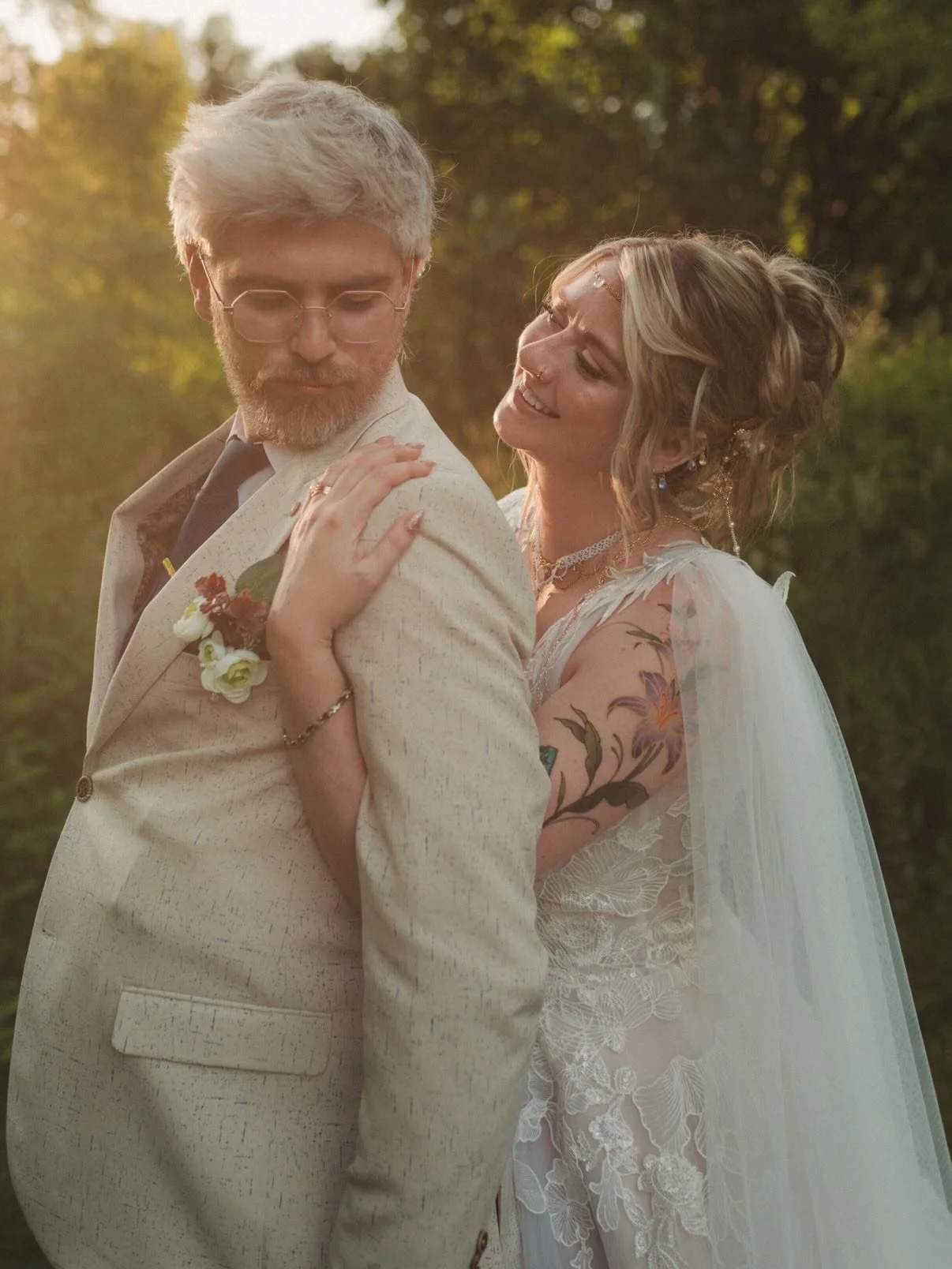 A couple dressed in vintage-style wedding attire, standing outdoors at sunset, with the woman smiling and looking lovingly at the man, who is looking down. The woman has tattoos on her arm and is wearing a lacy dress and jewelry. The man has gray hai