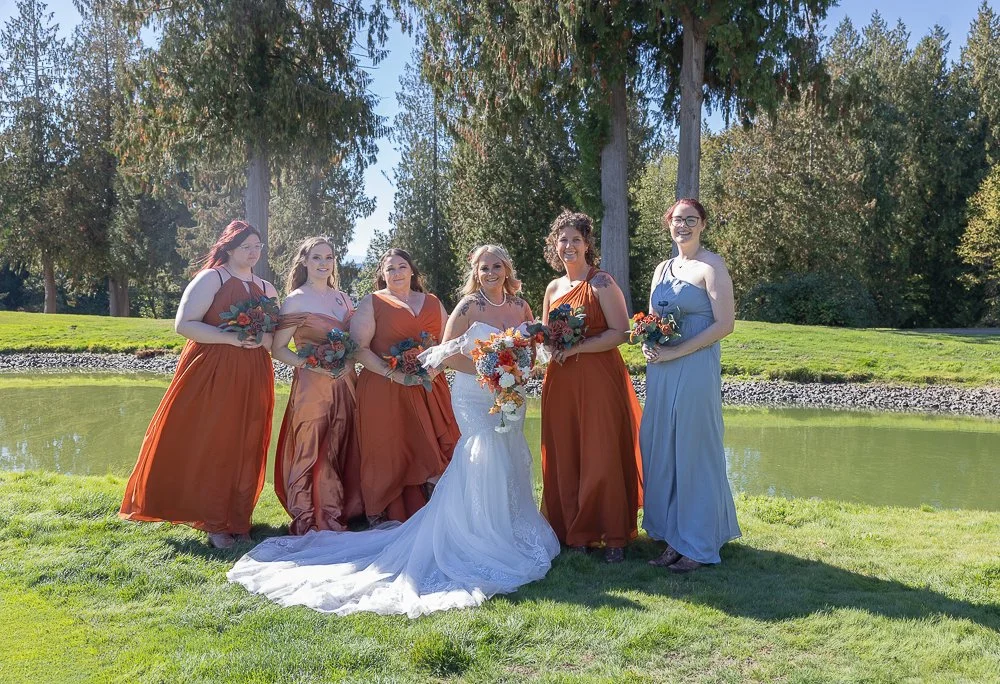 A bride in a white wedding dress standing with five bridesmaids in colorful dresses near a pond in a park.