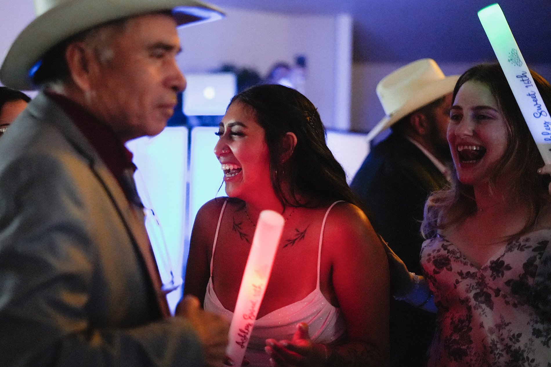 People celebrating at a party, two women smiling and laughing, wearing white cowboy hats, one holding a glow stick, vibrant party atmosphere.