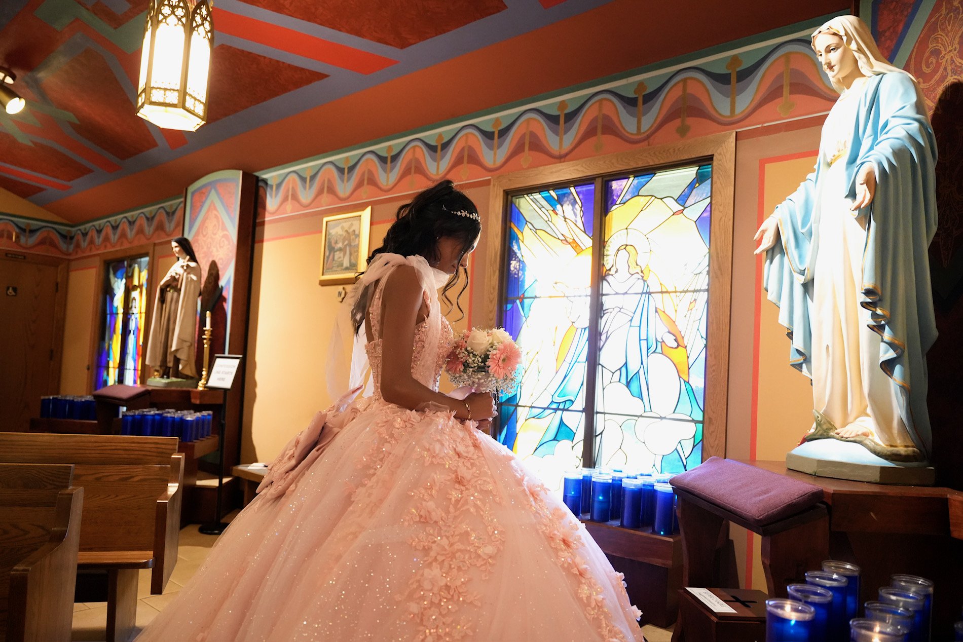 A young woman in a pink, elaborate ball gown holding a bouquet of flowers, standing inside a church near stained glass windows depicting religious imagery, with statues of religious figures in the background.