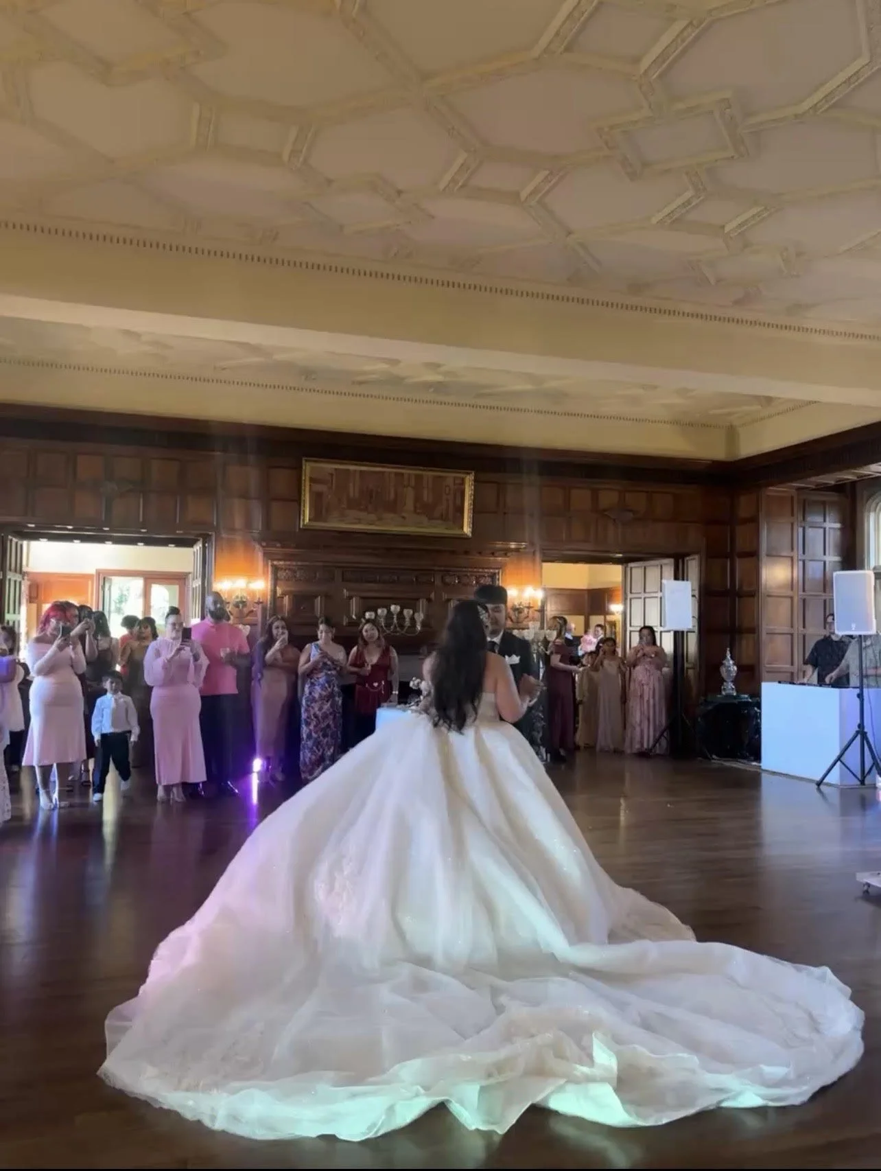 A bride and groom dance at their wedding reception as guests watch in a grand, wood-paneled ballroom.