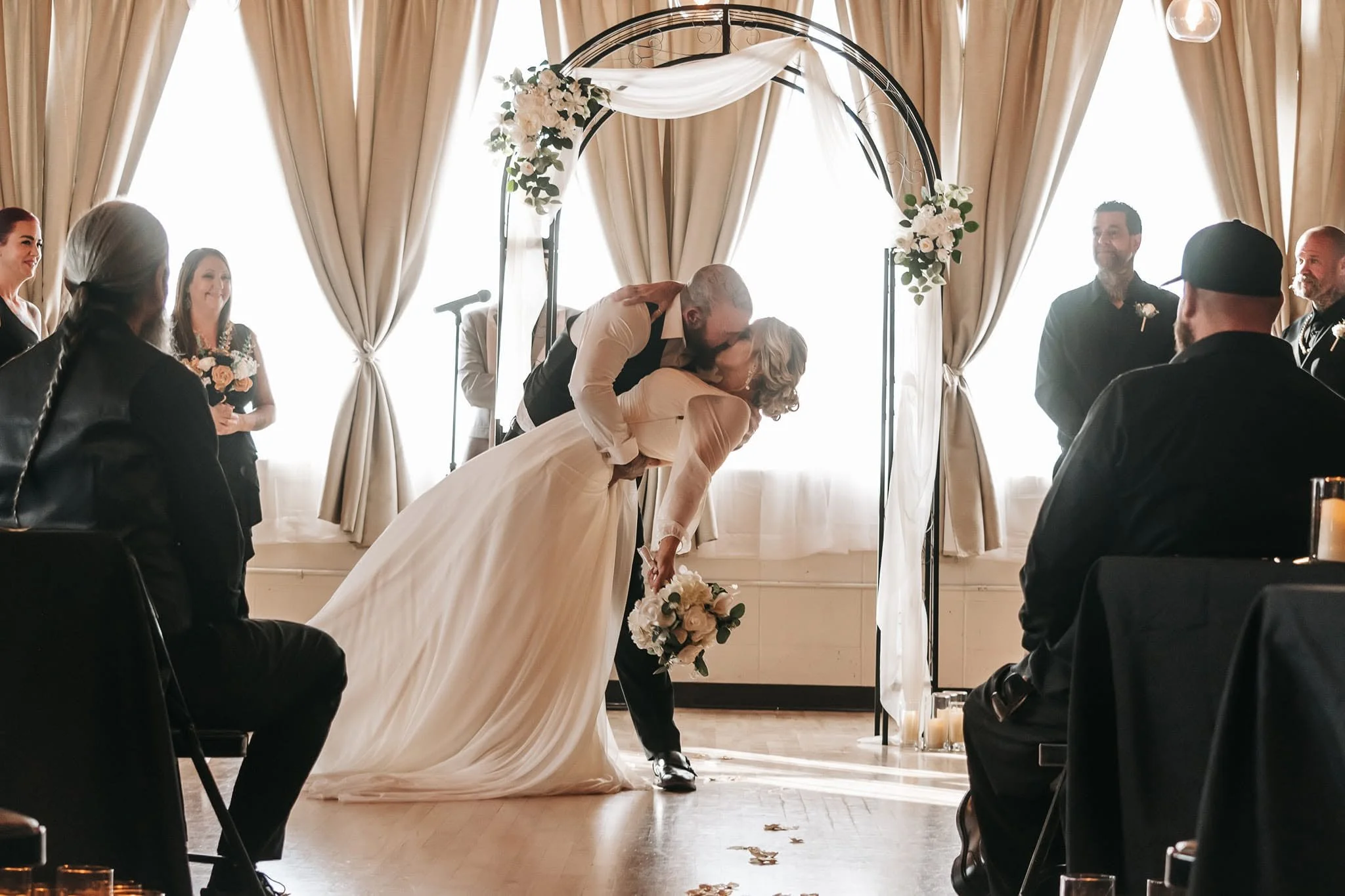 A bride and groom dancing during their wedding ceremony, with guests sitting and standing around in a decorated indoor venue.