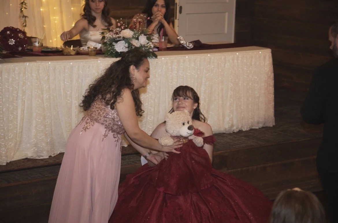 A girl in a red dress holding a teddy bear on her lap while an older girl in a pink dress helps her, at a celebration with a decorated table and floral centerpiece.
