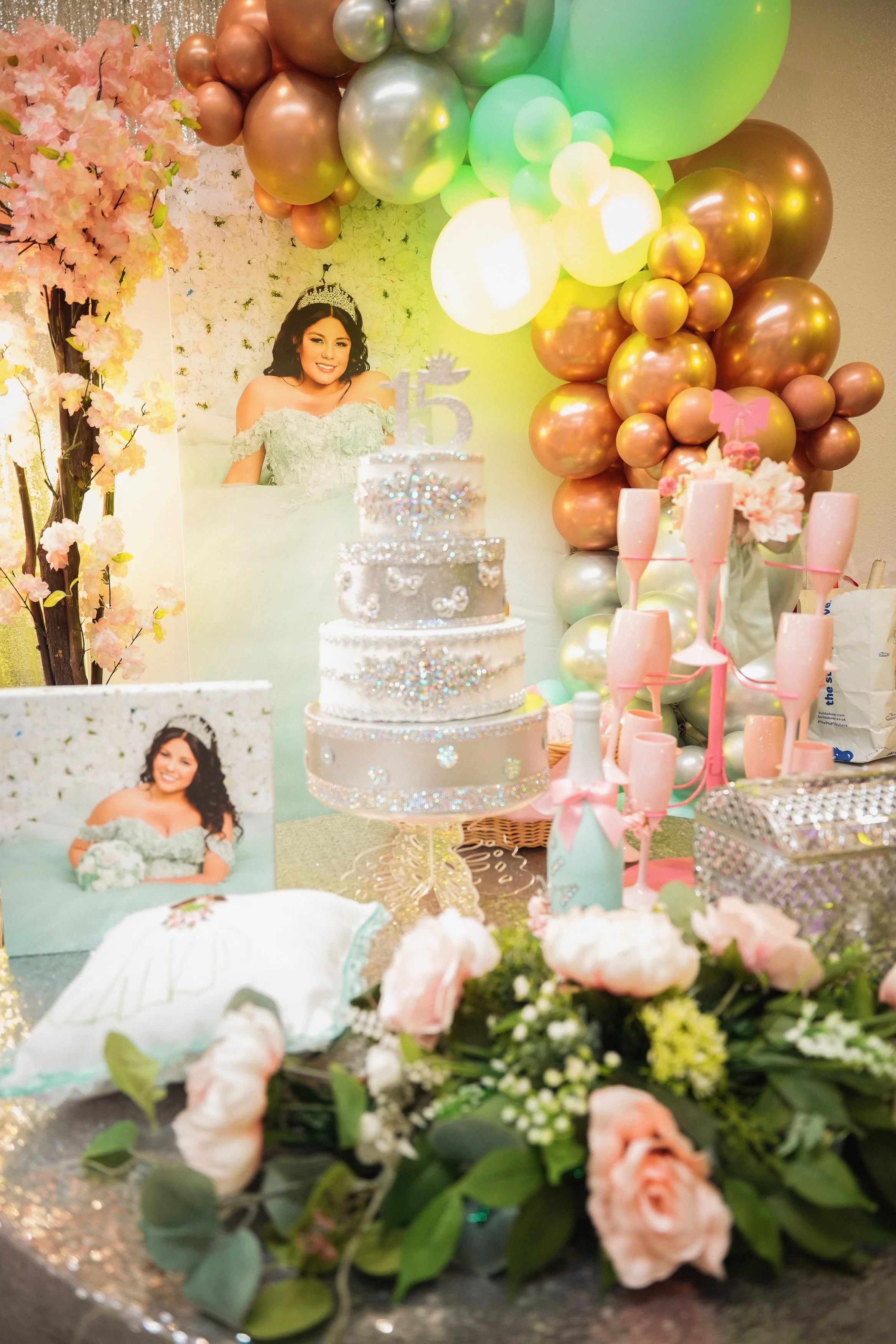 Decorative setup with pink flowers, balloons in shades of gold, green, and white, a birthday cake with the number 15, and photos of a woman, indicating a birthday celebration or sweet sixteen party.