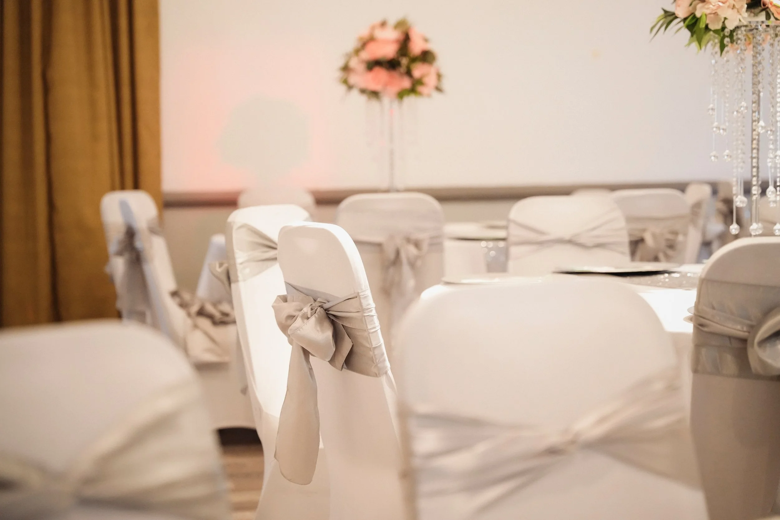 Close-up of white chairs decorated with bows in a decorated event hall with floral centerpieces and draped curtains.