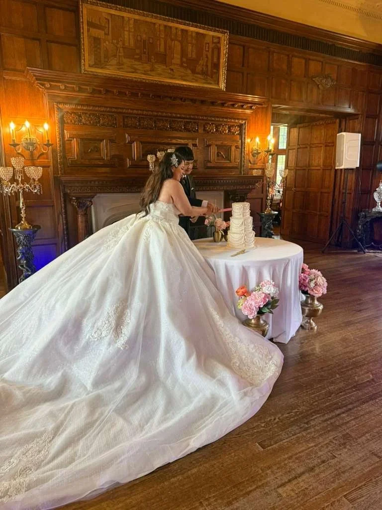 Couple in wedding attire cutting a wedding cake in a wood-paneled room with floral arrangements.