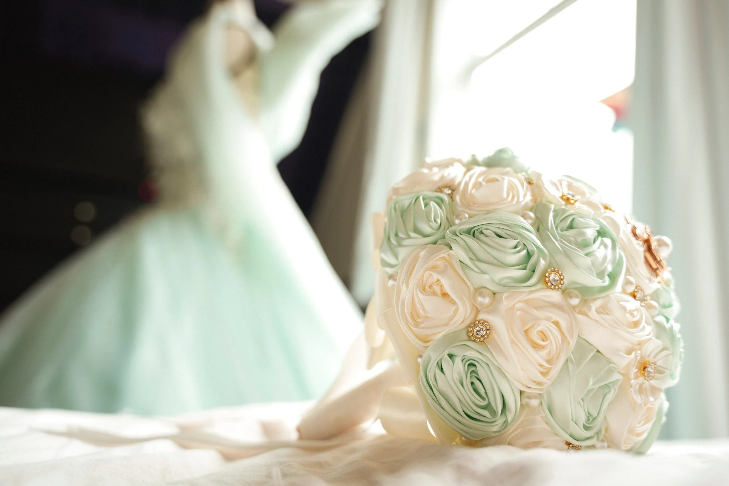 Close-up of a bouquet of cream, pale green, and peach-colored roses decorated with pearls and gold embellishments, resting on a white surface near a window with natural light shining through.