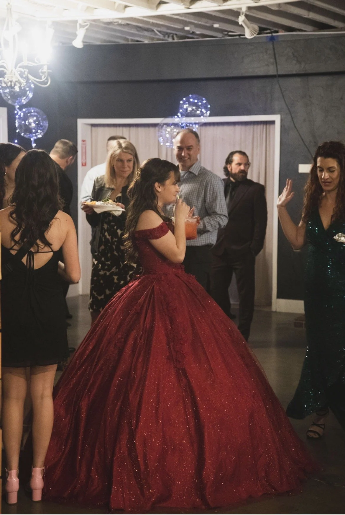 A woman in a red ball gown with a full skirt is at a party or celebration, standing with her back to the camera. She is sipping a drink while surrounded by other people, some holding drinks and plates, in a room decorated with blue lights and balloon