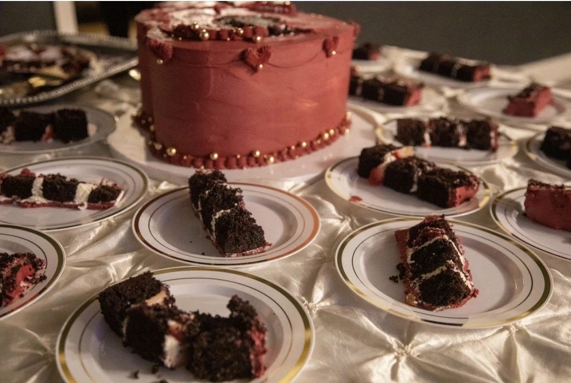 A red velvet cake with gold accents surrounded by slices of red velvet cake on decorative plates, set on a white tablecloth.