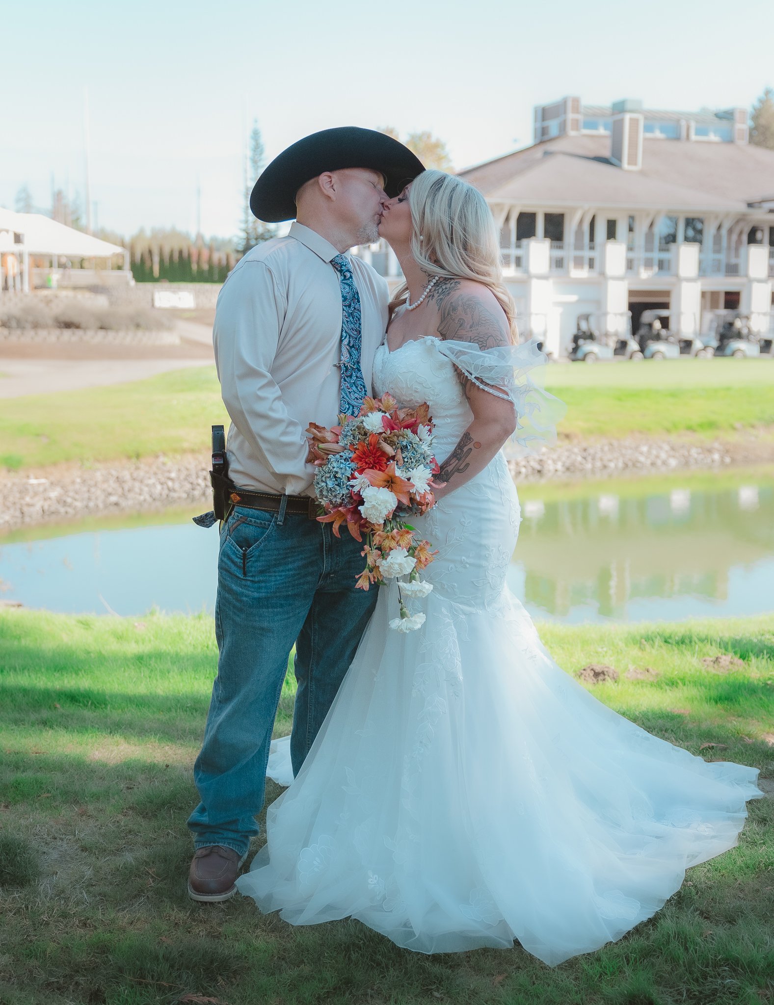 A couple kissing outdoors near a pond, with a large white house and golf carts in the background. The woman is in a white wedding dress holding a bouquet, and the man is in a white shirt, blue tie, and cowboy hat.