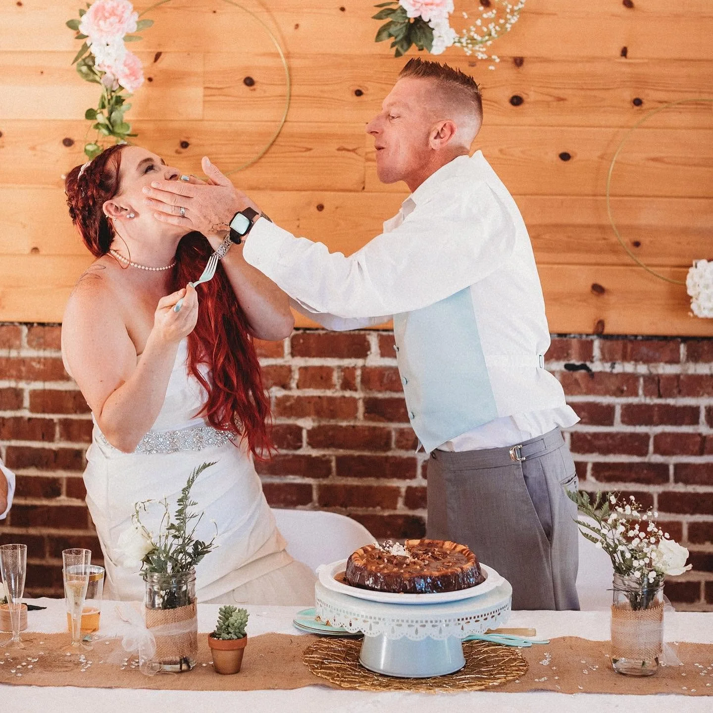 A bride and groom at wedding reception with a cake, floral decorations, and a brick wall background.