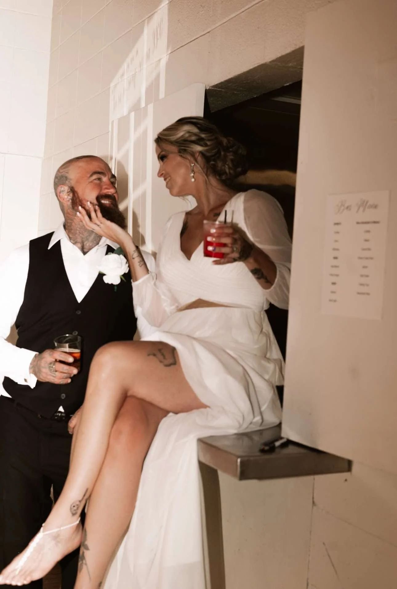 A man and a woman celebrating at a wedding, sitting close with the woman seated on the man's lap. The woman wears a white dress with a high slit, and the man is dressed in a black vest and white shirt. Both are smiling and holding drinks. The woman h