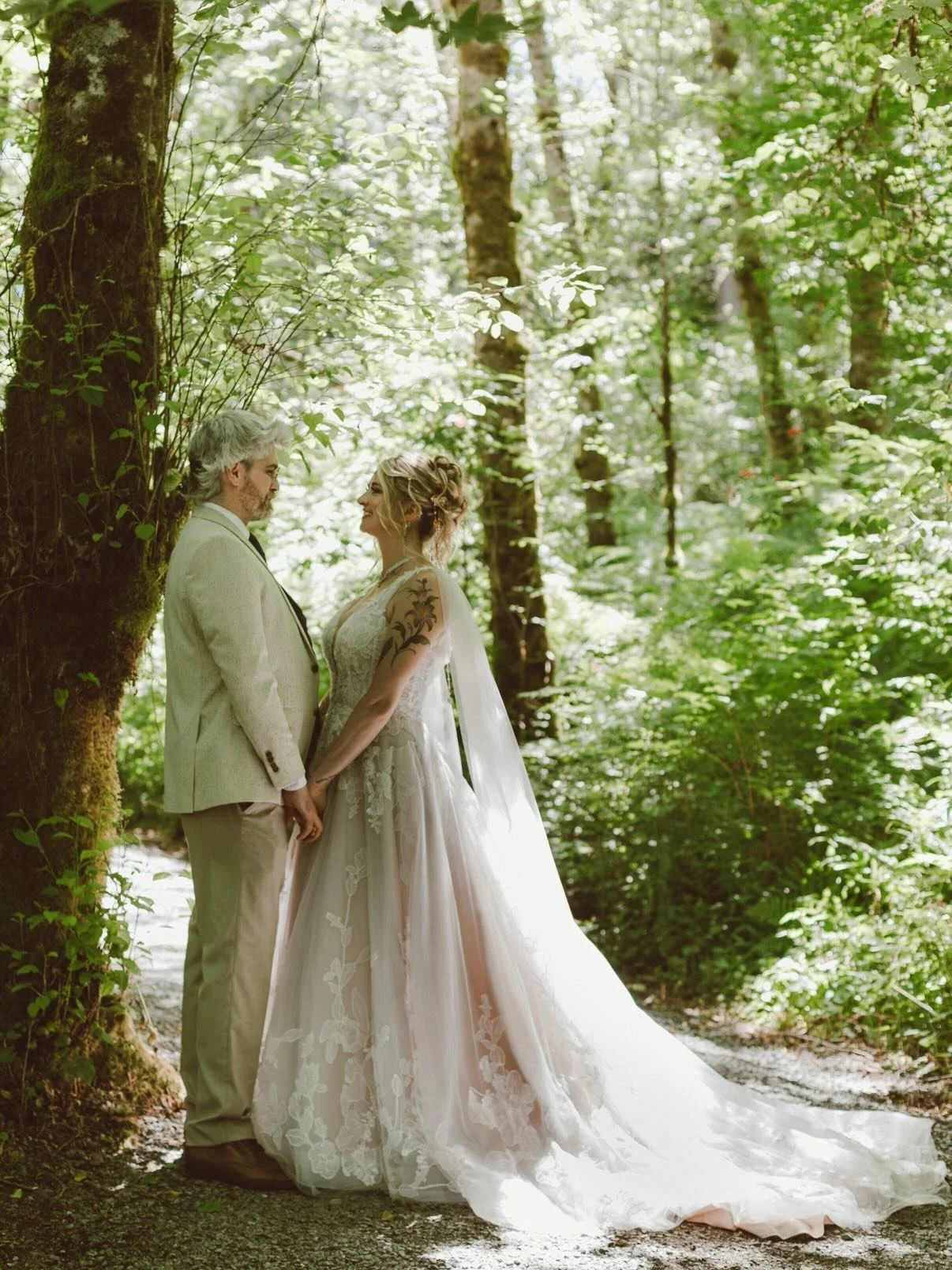 A couple dressed in wedding attire, standing close and holding hands in a lush green forest, gazing at each other.