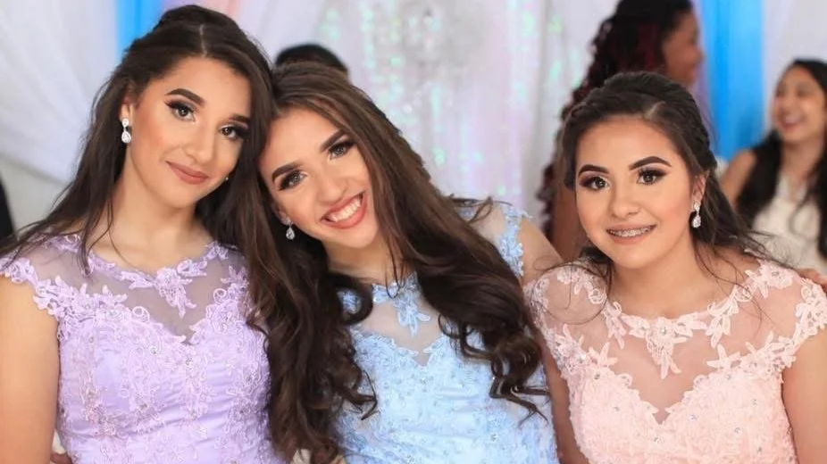 Three young women wearing pastel-colored lace dresses, posing at a festive event, with other smiling women in the background.