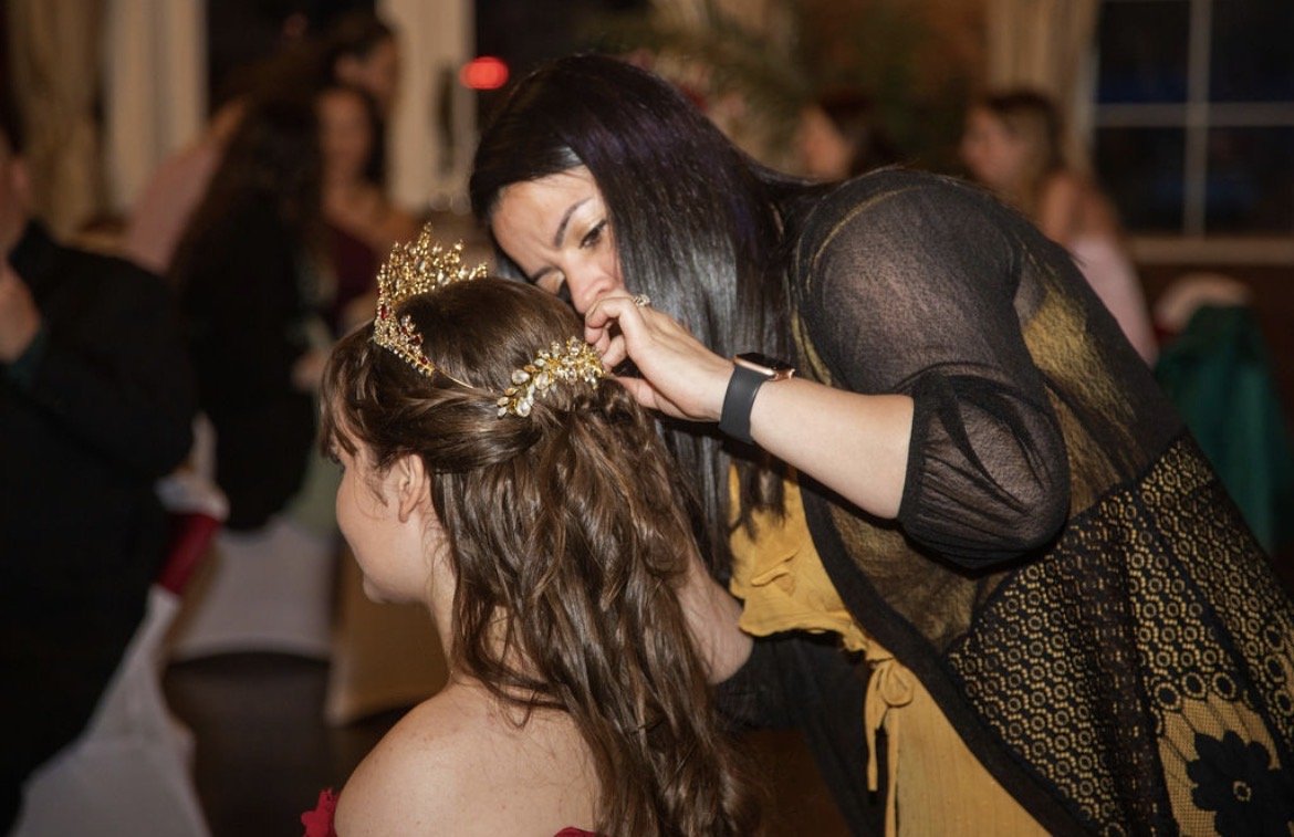 A woman helping a young girl with her hair and makeup at a social event, with other people in the background.