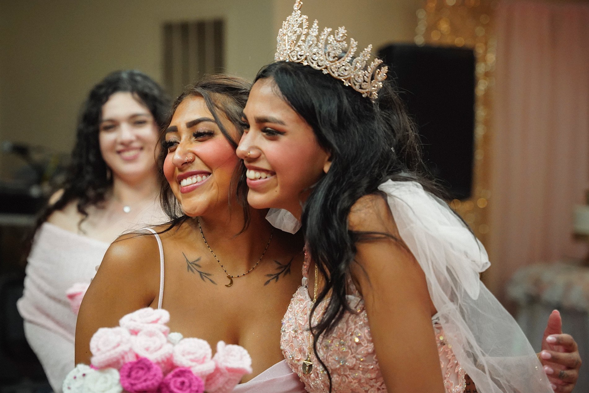 Celebration scene with three women, one wearing a crown, smiling and embracing, with pink and white floral decorations in the foreground.