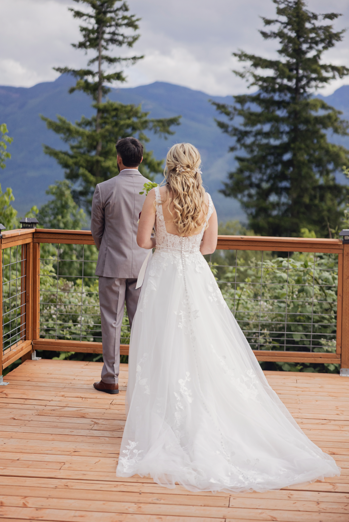 A bride and groom stand on a wooden balcony overlooking a lush green landscape and mountains in the distance, with the bride in a white wedding gown and the groom in a gray suit.